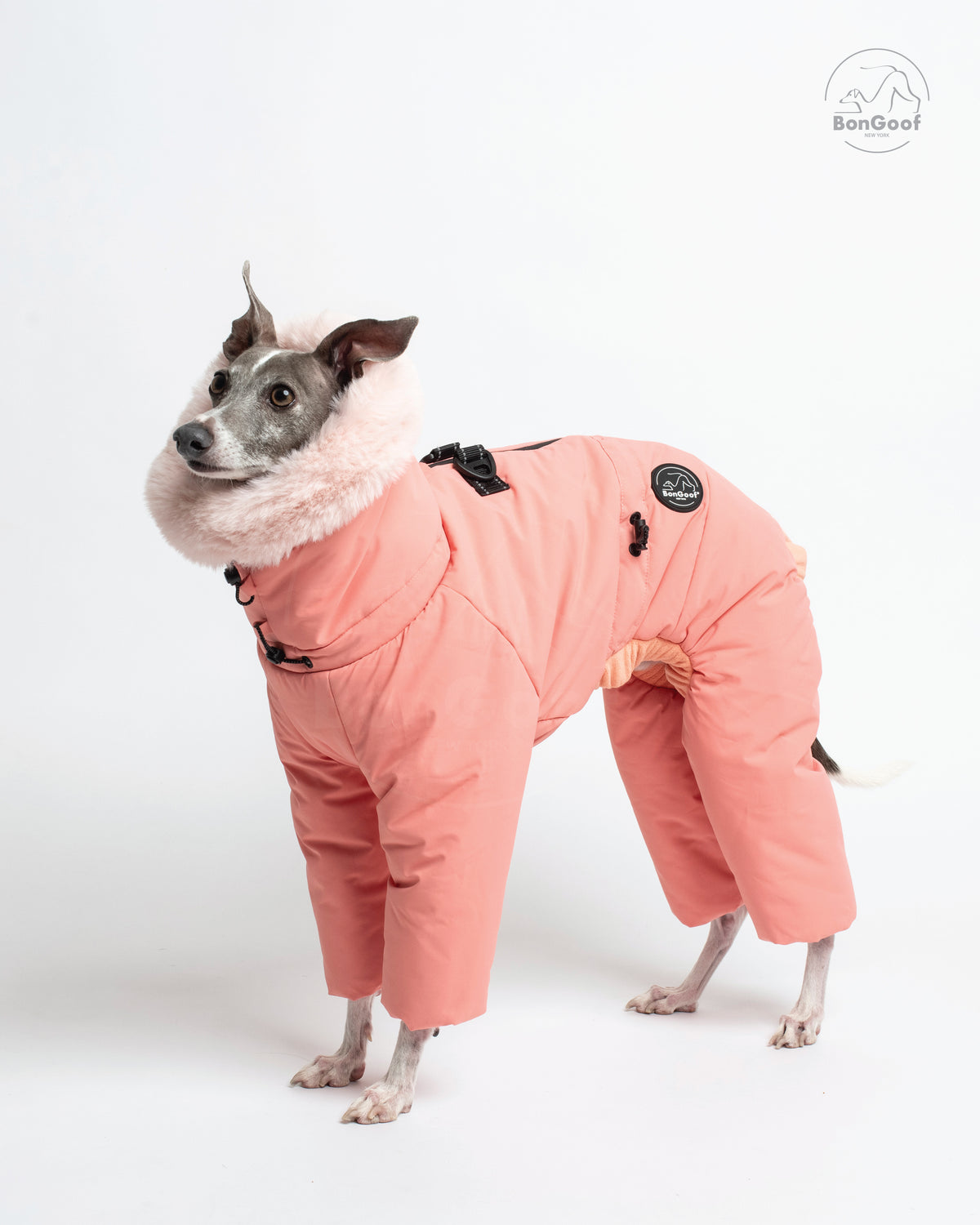 Dog wearing the BonGoof SUIT-PREMO snowsuit in Peach, side view showing the peach-colored body, covered legs, faux-fur-trimmed hood and logo patch.
